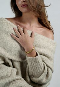 Maglione beige lavorato a maglia con design off-shoulder, con una mano appoggiata sul petto. Un braccialetto di metallo dorato con texture è indossato al polso.