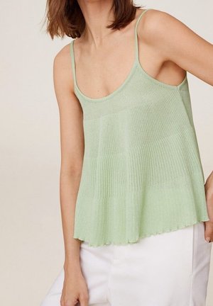 Woman wearing a light green knit sleeveless top with spaghetti straps and white pants against neutral background.