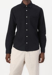 Sort langærmet skjorte af letvægtsstof med knappet krave, brystlomme og hvide knapper, kombineret med lyse bukser.
