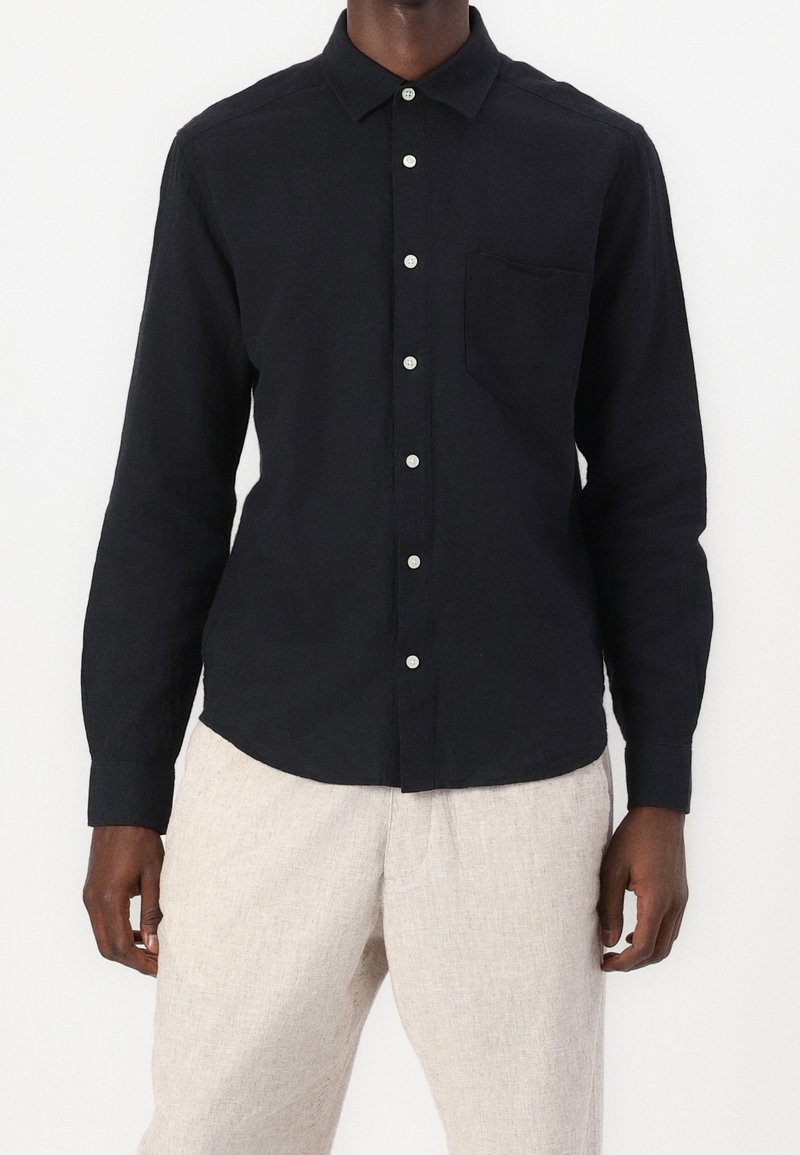 Sort langærmet skjorte af letvægtsstof med knappet krave, brystlomme og hvide knapper, kombineret med lyse bukser.