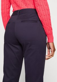 Žena v navy modrých upnutých kalhotách a jasně korálově zbarveném svetr s pleteným vzorem, ruka uvolněná po boku, zobrazená zezadu ve výšce pasu.