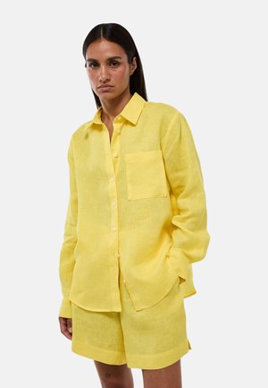 Femme portant une chemise jaune ample à manches longues boutonnée avec une poche poitrine et un short jaune assorti sur un fond uni.