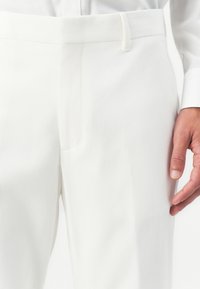 Artimiausias žmogaus kadrą, vilkinčio pritaikytas baltas kelnes su diržo kilpelėmis, šoniniu kišenė ir matoma balta marškinėlio rankovė dešinėje.