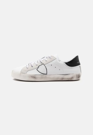 Basket basse blanche avec des accents en daim gris, empiècement noir au talon, logo en forme de bouclier sur le côté, lacets imprimés et semelle usée à l’aspect sale.