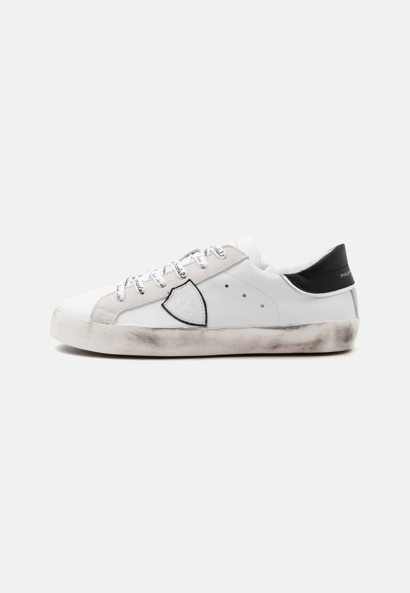 Basket basse blanche avec des accents en daim gris, empiècement noir au talon, logo en forme de bouclier sur le côté, lacets imprimés et semelle usée à l’aspect sale.