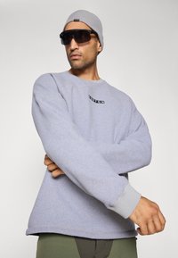 Jasnoszara bluza z miękką teksturą, z prążkowanymi mankietami i czarnym logo na przodzie. Noszona z szarym kaszkietem i ciemnymi okularami przeciwsłonecznymi.
