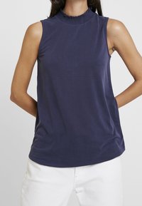 Top sin mangas de color azul marino con cuello alto y detalles de volantes. Fabricado en tejido suave con una textura lisa y un corte relajado.