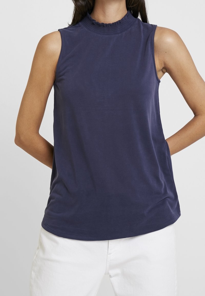 Top sin mangas de color azul marino con cuello alto y detalles de volantes. Fabricado en tejido suave con una textura lisa y un corte relajado.
