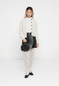 Vrouw in een witte cardigan, zwarte leren rok, witte panty's, zwarte flats, met een kleine zwarte handtas, staande tegen een effen witte achtergrond.
