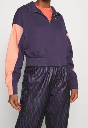 Person iført en lilla og ferskenfarvet Nike sweatshirt med halv lynlås, elastisk talje og skinnende lilla bukser med zebrastriber.