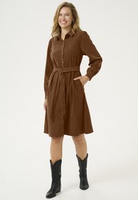 Robe chemise en velours côtelé marron avec des manches longues, patte de boutonnage à l'avant et ceinture à la taille. Comprend deux poches latérales et est assortie à des bottes de cowboy noires.