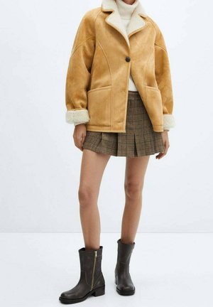 Femme portant une veste en shearling beige, un col roulé blanc, une mini-jupe plissée marron à carreaux et des bottes noires mi-mollet, debout devant un fond blanc.