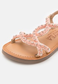Sandalia de punta abierta con un upper de cuentas rosa claro y acentos de strass, una plantilla de cuero marrón y una suela texturizada marrón.