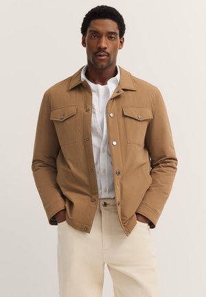 Braune Bomberjacke mit zwei Brusttaschen, silbernen Druckknöpfen und glatter Textur, getragen über einem weißen Hemd und beigen Hosen.