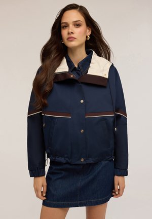 Marineblaue Jacke mit cremefarbenem Kragen und braunen Akzenten, ausgestattet mit einem Reißverschluss, einem Kordelzugsaum und Fronttaschen, getragen über einem denims Rock.