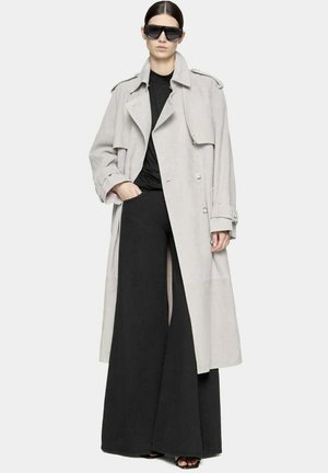 Trench coat grigio chiaro con design doppiopetto, ampi revers e bottoni grandi, abbinato a pantaloni larghi neri e occhiali da sole.