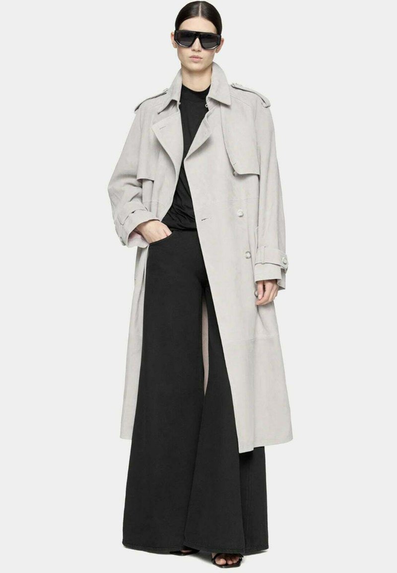 Trench coat grigio chiaro con design doppiopetto, ampi revers e bottoni grandi, abbinato a pantaloni larghi neri e occhiali da sole.
