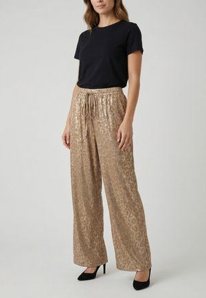 Femme portant un haut noir à manches courtes, un pantalon ample taille haute imprimé léopard doré avec cordon, et des talons hauts noirs, debout sur fond gris.