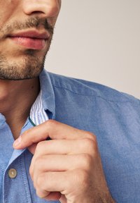 Šviesiai mėlynas marškinis su mygtukų apykakle, kuriame yra balta ir mėlyna dryžuota vidinė danga; rankos, reguliuojančios apykaklę.