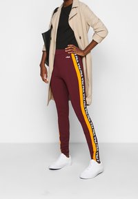 Vinfärgade leggings med svarta och gula sidostråk samt vit FILA-logga, kombinerat med en svart topp och en beige trenchcoat, samt vita sneakers.