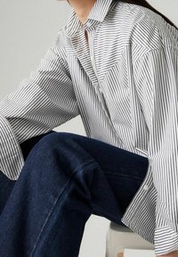 Chemise à rayures blanche et noire à boutons, coupe décontractée, manches larges et col pointu. Associée à un pantalon en denim foncé.