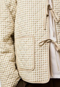 Veste matelassée en vichy beige et blanc avec une grande poche appliquée et des fermetures à ruban nouées, portée sur une chemise blanche.