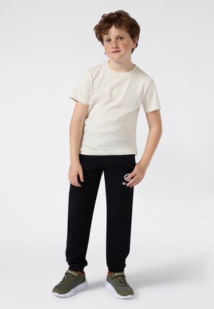 T-shirt à manches courtes beige clair avec logo brodé, associé à un pantalon de survêtement noir avec impression de logo. Des baskets vert olive complètent le look.