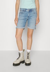 Šviesiai mėlyni džinsiniai šortai su sulenktais kraštais, derinami su smėlio spalvos raišteliais bateliais, turinčiais storą juodą padą ir juodais raišteliais, dėvimi su baltais kojiniais.