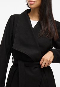 Abrigo negro con cuello ancho, cinturón y textura suave. Presenta un diseño de escote en V profundo y mangas largas, mostrado sobre un fondo neutral.