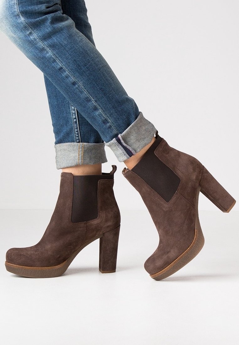 Bottines en daim marron à talons épais portées avec un jean bleu retroussé sur un fond clair uni.