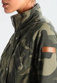 Armádní zelený maskovací kabát s vysokým límcem, patentními knoflíky, čtyřmi předními kapsami a hnědým koženým logem značky na rameni.