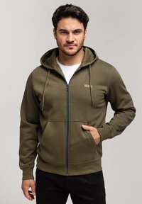 Oliwkowa bluza z kapturem na zamek błyskawiczny, z kieszeniami z przodu, ściągaczami przy mankietach i u dołu. Posiada małe logo na piersi oraz gładką teksturę materiału.