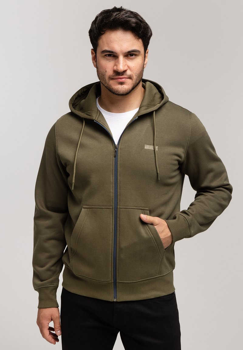 Oliwkowa bluza z kapturem na zamek błyskawiczny, z kieszeniami z przodu, ściągaczami przy mankietach i u dołu. Posiada małe logo na piersi oraz gładką teksturę materiału.
