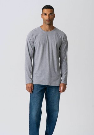 Mężczyzna stojący przodem, ubrany w prostą szarą bluzkę z długim rękawem i niebieskie dżinsy na jednolitym jasnym tle.