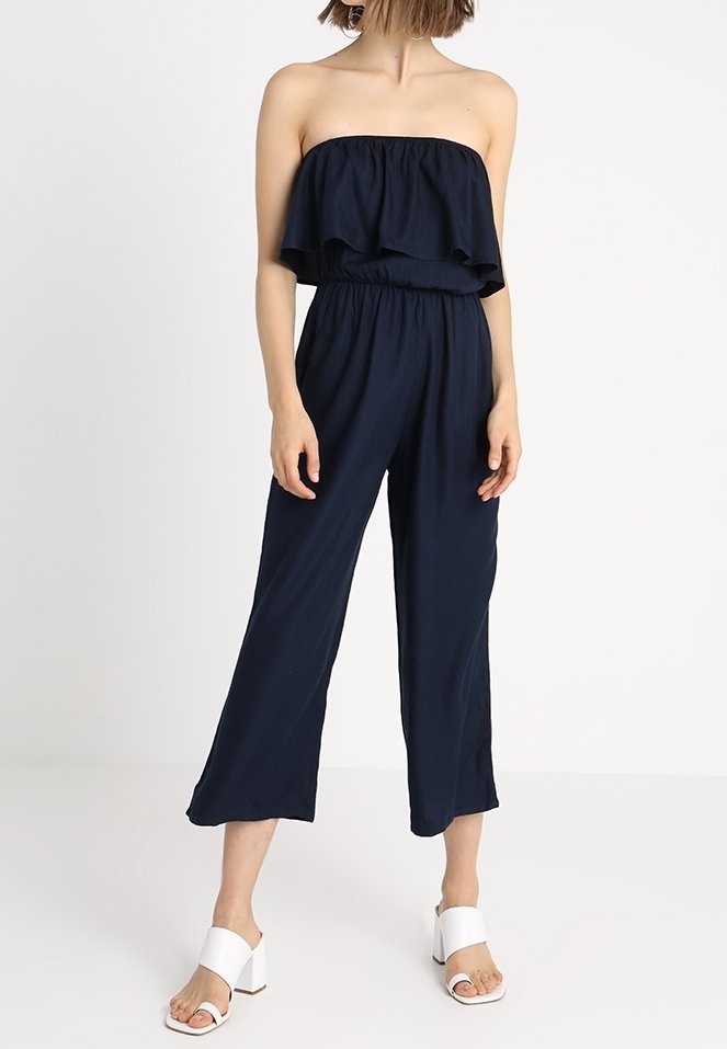 Námořnický modrý overal bez ramínek s volánkovým vrškem, elastickým páskem v pase a širokými nohavicemi. Modelka má na sobě bílé sandály na blokovém podpatku.