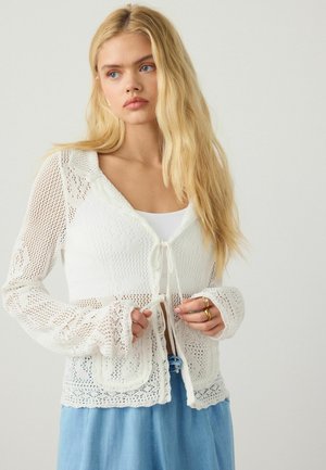 Joven mujer con cabello rubio largo que lleva un cárdigan blanco de punto anudado al frente y pantalones azul claro sobre un fondo liso.