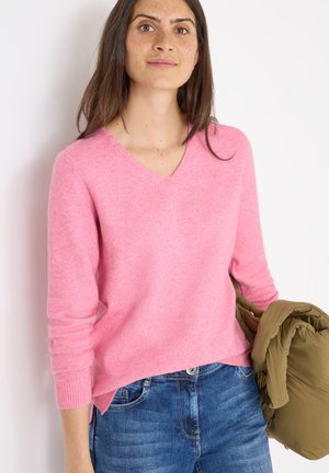 Rosa V-Ausschnitt pullover mit langen Ärmeln, aus weichem Material. Pauliert mit blauen Jeans und hält eine olivgrüne Daunenjacke.