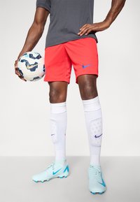 Røde Nike shorts, blåt logo, hvide strømper med skinnebeskyttere, og lyseblå fodboldstøvler med blå detaljer. Holder en mønstret fodbold.