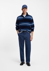 Polaire à rayures bleu marine et bleu clair avec col, associée à un jean denim foncé ample et des mocassins noirs, complétée par une casquette bleue.