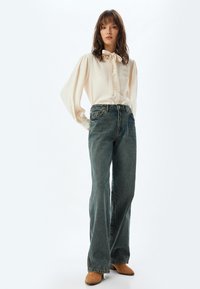 Blusa crema con un fiocco al collo, abbinata a jeans in denim scuro a vita alta e a gamba larga. Texture morbida, vestibilità comoda e chiusura con bottoni.