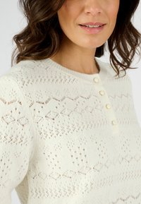 Pull en maille blanc cassé avec un motif texturé. Il présente un col rond et quatre petits boutons ronds sur le devant.