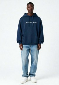 Marineblauwe hoodie met witte tekst "IK HEB GEZEGD DAT, OVER JOU," losse pasvorm, kangoeroezak, gecombineerd met lichtblauwe jeans en witte sneakers.