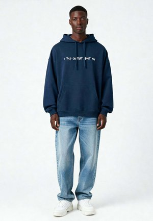 Felpa navy con testo bianco che recita "TI HO DETTO DI QUELLO, RIGUARDO A TE," vestibilità comoda, tasca a marsupio, abbinata a jeans azzurri chiari e sneakers bianche.