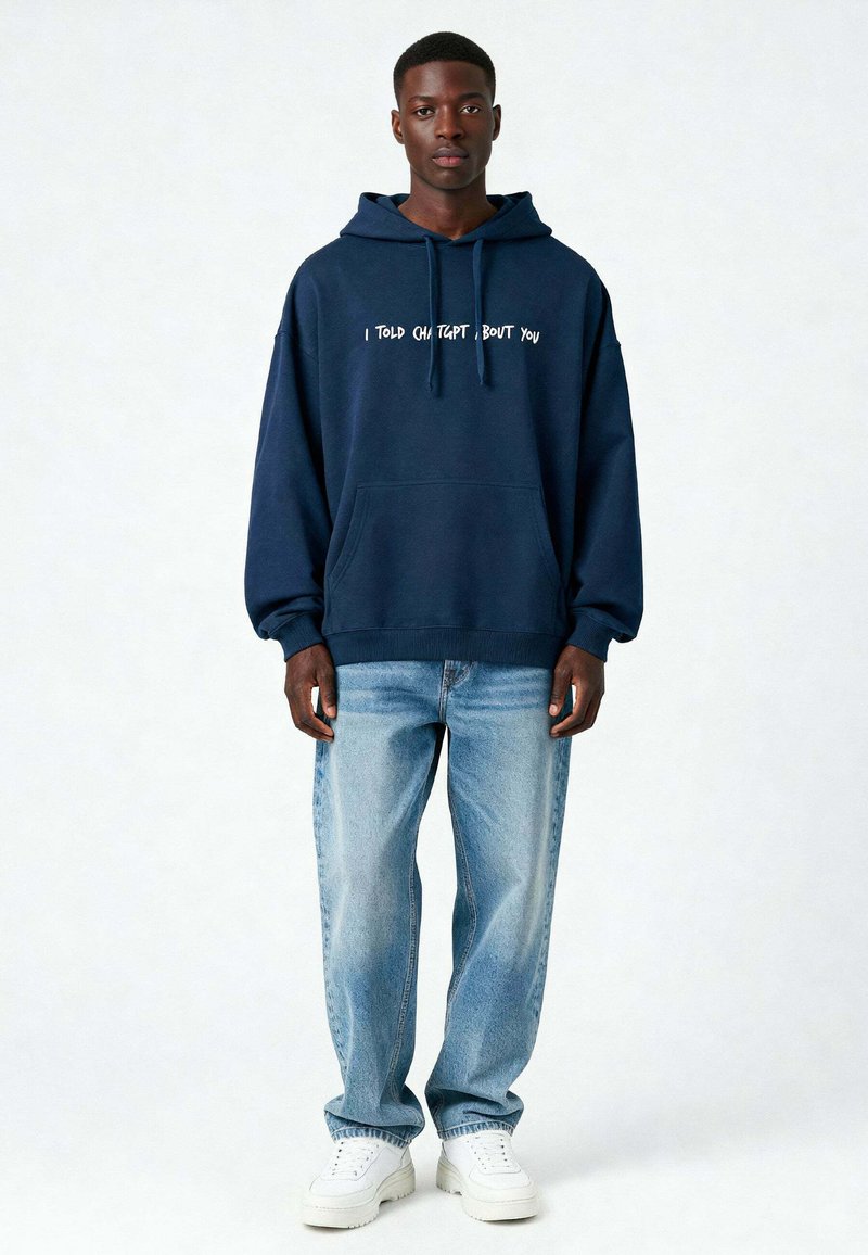 Marineblauwe hoodie met witte tekst "IK HEB GEZEGD DAT, OVER JOU," losse pasvorm, kangoeroezak, gecombineerd met lichtblauwe jeans en witte sneakers.