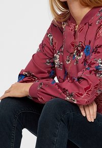 Veste bomber zippée à motifs floraux en tissu bordeaux avec des motifs floraux bleus et crème, poignets côtelés, et une texture lisse.