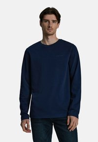 Granatowa bawełniana koszula z długim rękawem, z okrągłym dekoltem i subtelnym logo na piersi, w połączeniu z ciemnymi jeansami. Gładka faktura i luźny krój.