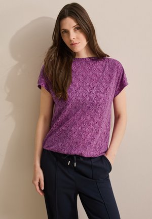 Top morado de manga corta con un patrón de hojas, cuello redondo y textura suave; combinado con pantalones oscuros, el modelo se encuentra de pie contra un fondo neutro.
