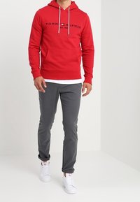 Sudadera roja con capucha y bolsillo frontal, con el logo negro "TOMMY HILFIGER", combinada con pantalones grises y zapatillas blancas.