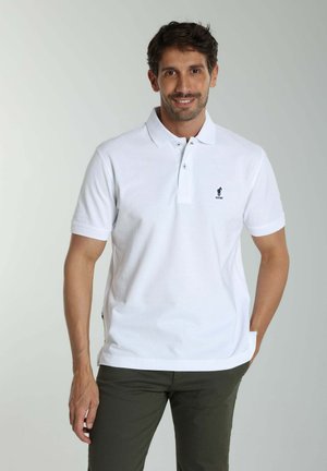 Homme souriant portant un polo blanc à manches courtes avec un petit logo foncé sur la poitrine et un pantalon vert foncé, debout devant un fond neutre.