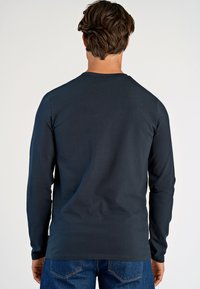 T-paita tummansinisestä puuvillasta, pitkähihaisena, jossa on pyöreä kaula-aukko ja suora leikkaus. Näkyvät ompeleet ja pieni tuotemerkki sivussa.